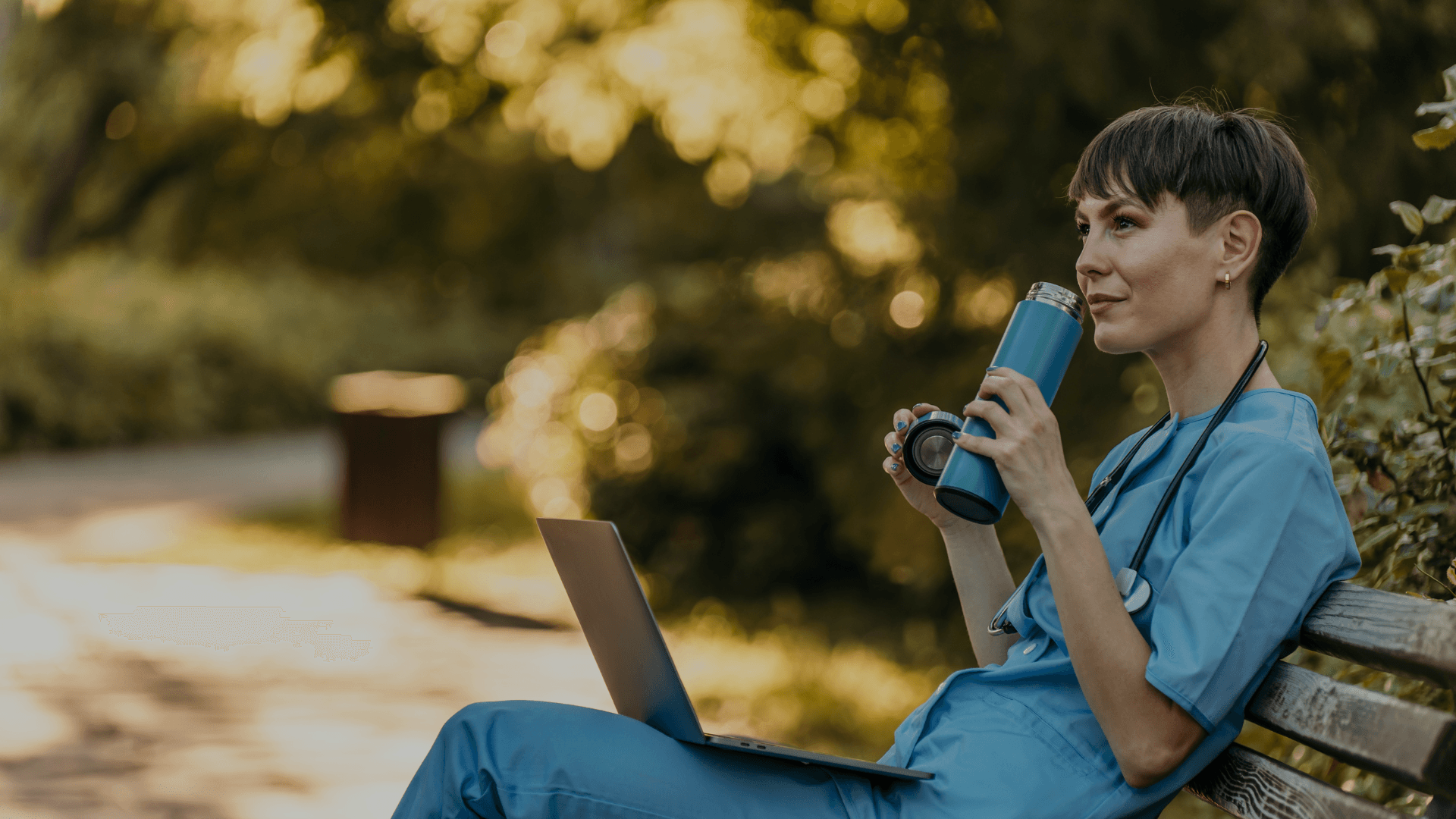 Zorgverlener in blauw zorgtuniek zit op een parkbankje in de pauze met een laptop, houdt een thermosbeker vast