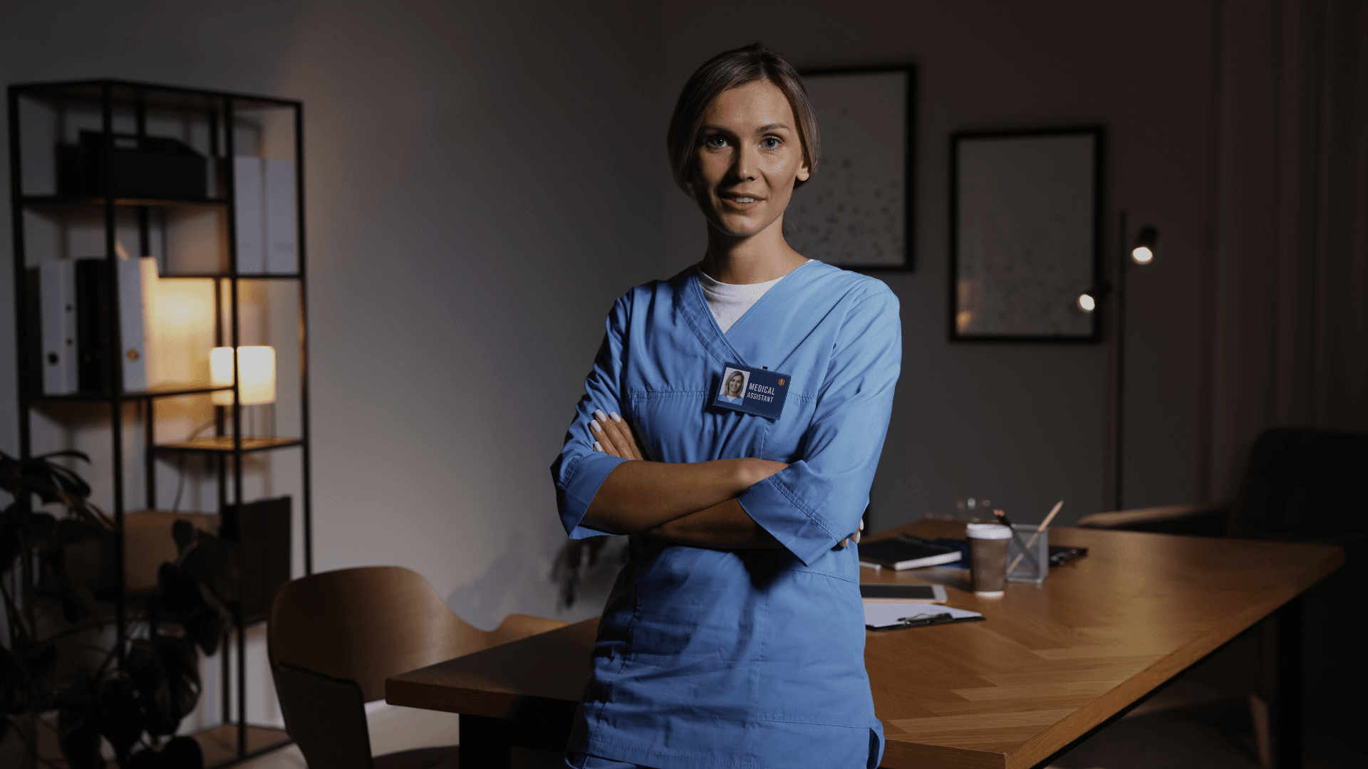 Vrouw in blauw zorgtuniek staat zelfverzekerd met gekruiste armen in een kantoor in de avond
