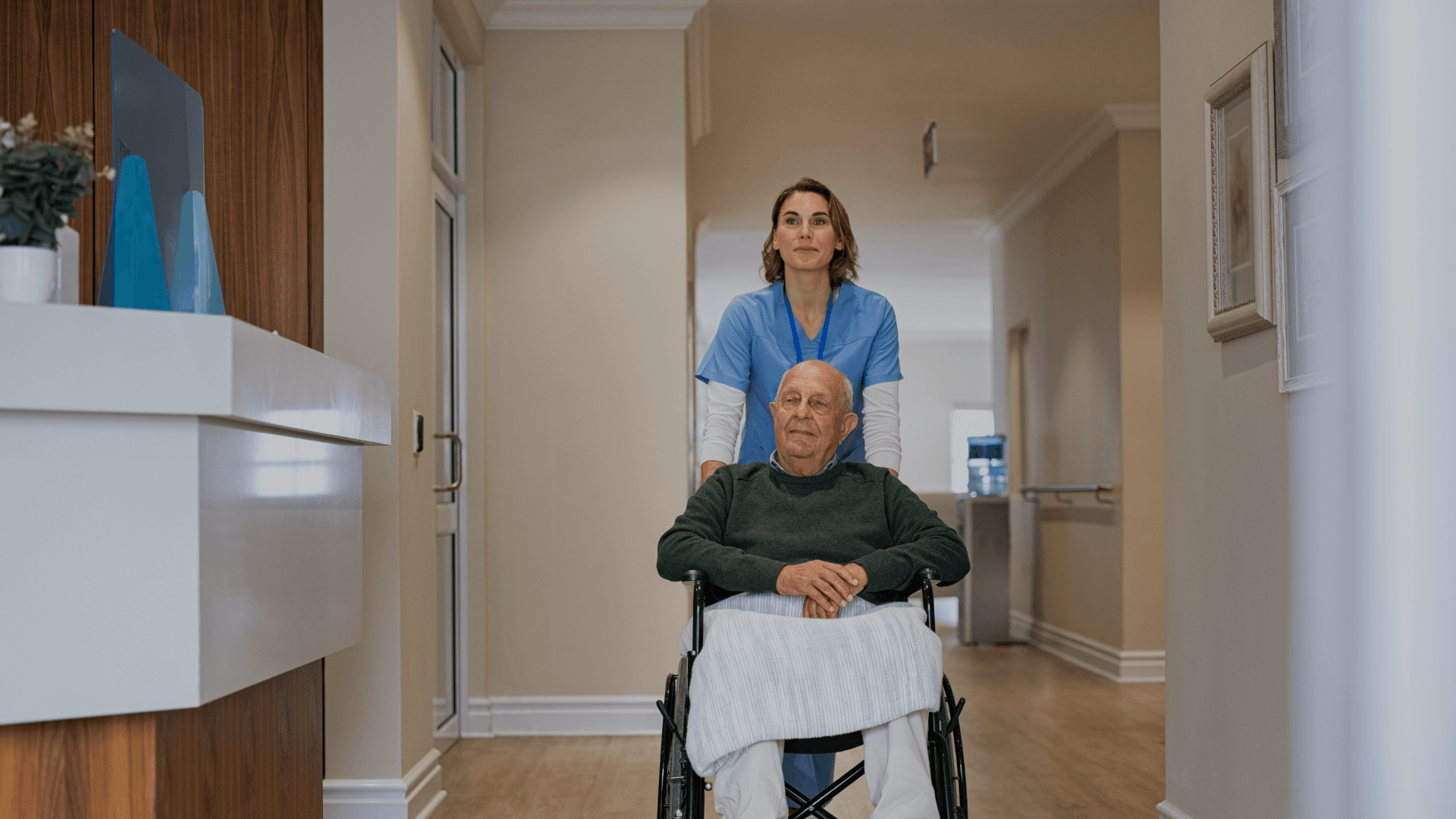 Verpleegkundige in blauw uniform duwt een oudere man in een rolstoel door een gang, met een deken over zijn schoot