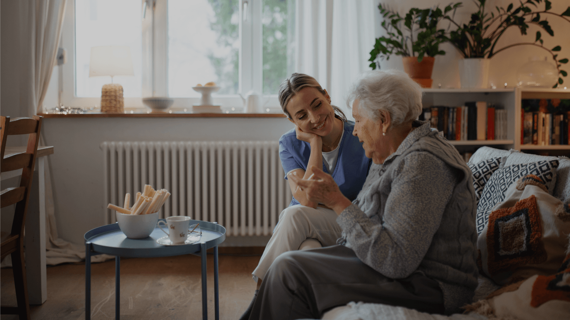 Verzorgende in blauwe zorgtuniek luistert aandachtig naar een oudere vrouw in een gezellige kamer