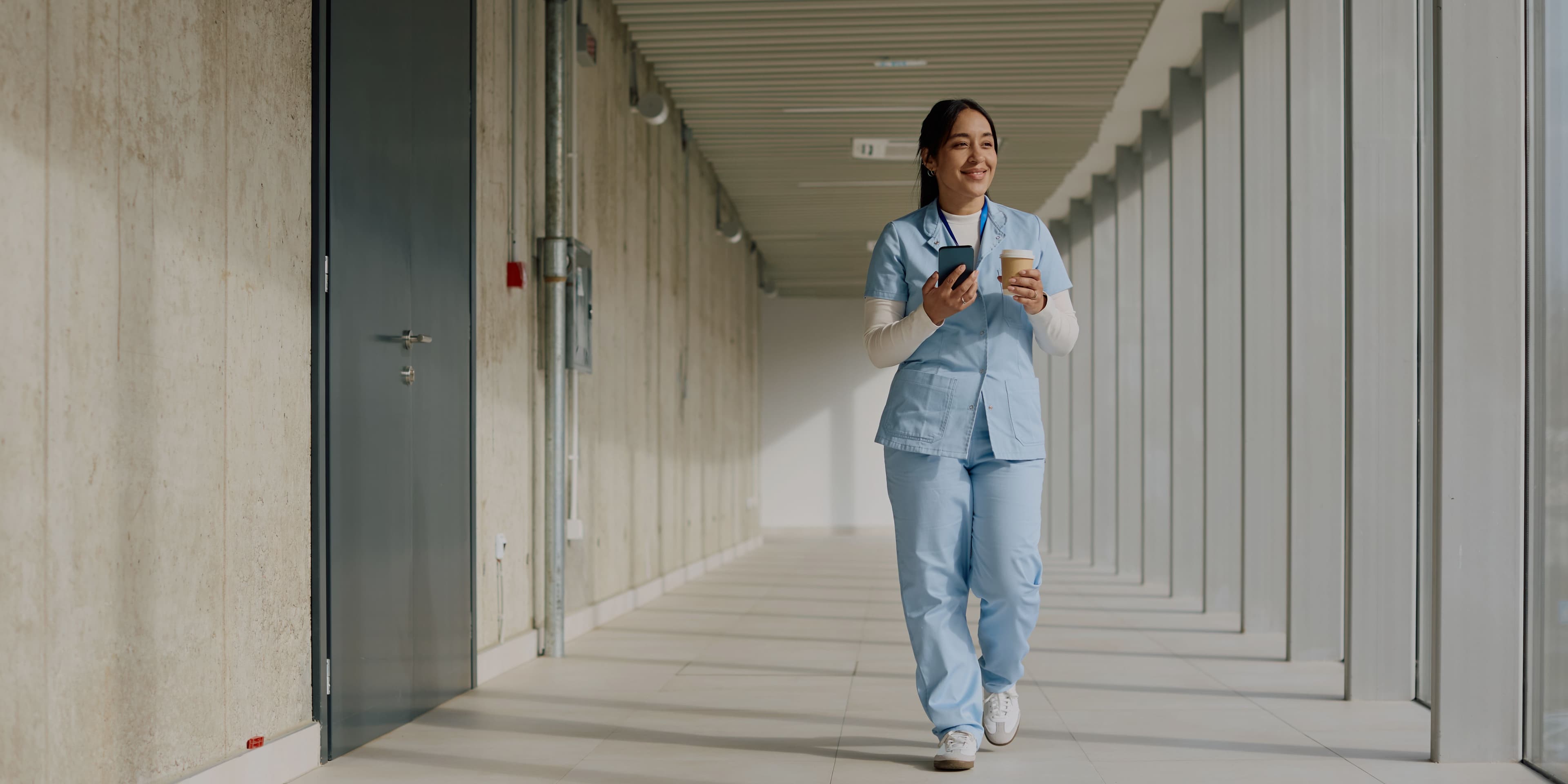 Verpleegkundige loopt door een lichte gang met koffie en telefoon tijdens een werkdag in de zorg