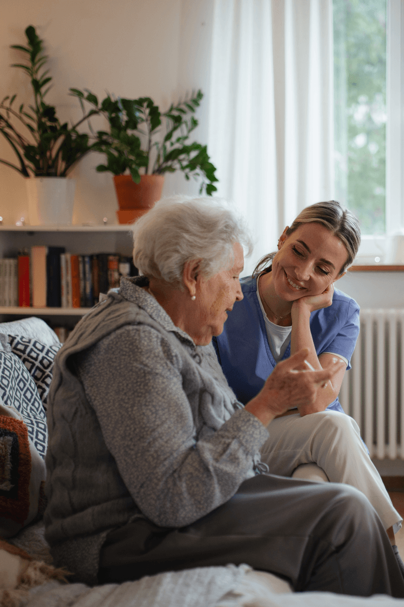 Verzorgende in blauwe zorgtuniek luistert aandachtig naar een oudere vrouw in een gezellige kamer