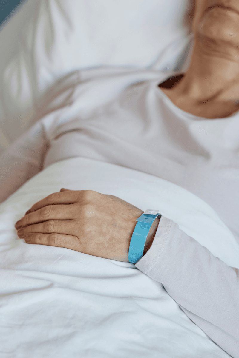 Persoon rust in een ziekenhuisbed met een blauw polsbandje en handen op een witte deken